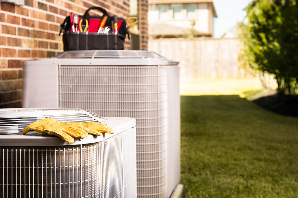 Bag of repairman's work tools, gloves on top of air conditioner units outside a brick home. Service industry, working class.